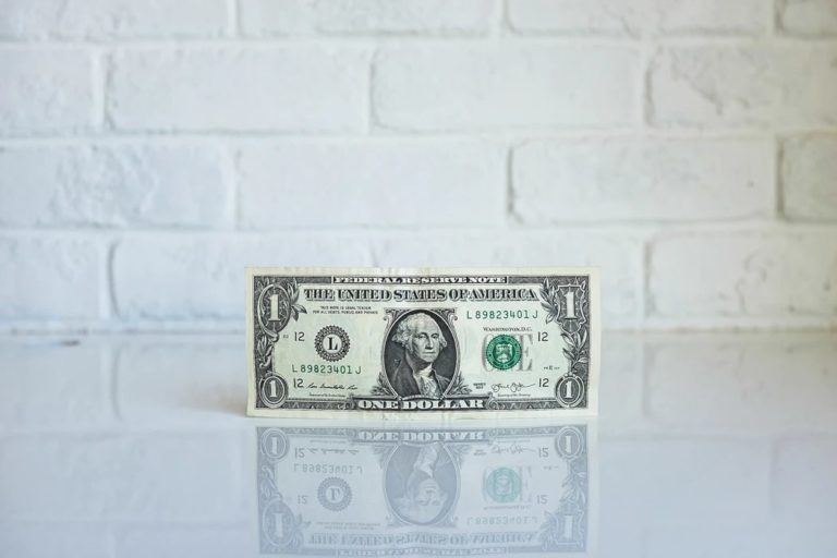 A one-dollar bill stands on a white countertop in front of a white brick wall