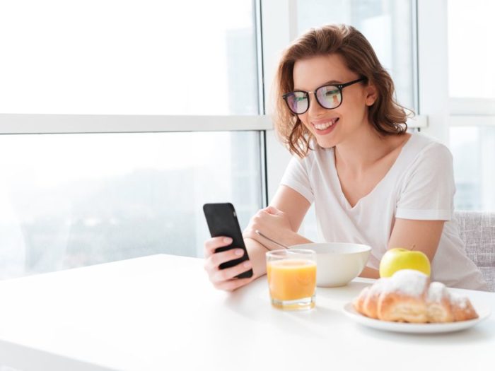 A happy young woman eats breakfast while looking at her phone