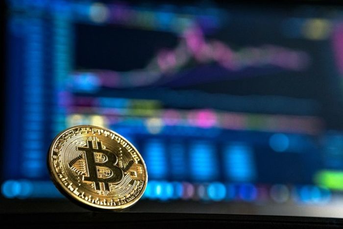 A bitcoin sits in front of a computer screen showing the value of the cryptocurrency rising
