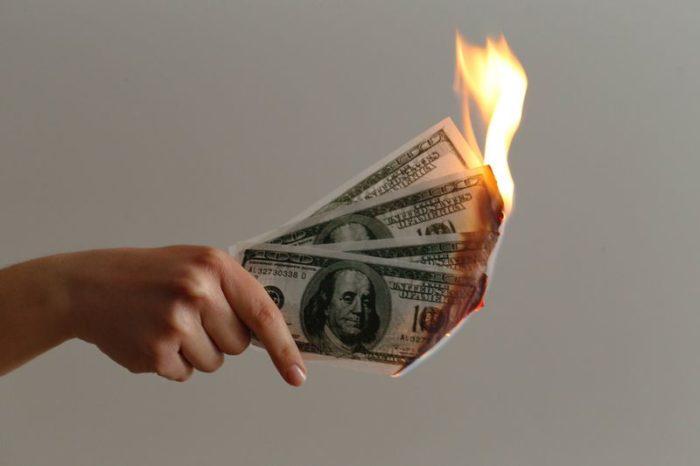 A woman holds out a wad of one-hundred-dollar bills on fire