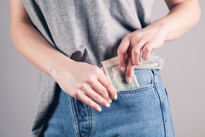 A woman wearing a grey shirt pulls out some paper money from the pocket of her blue jeans